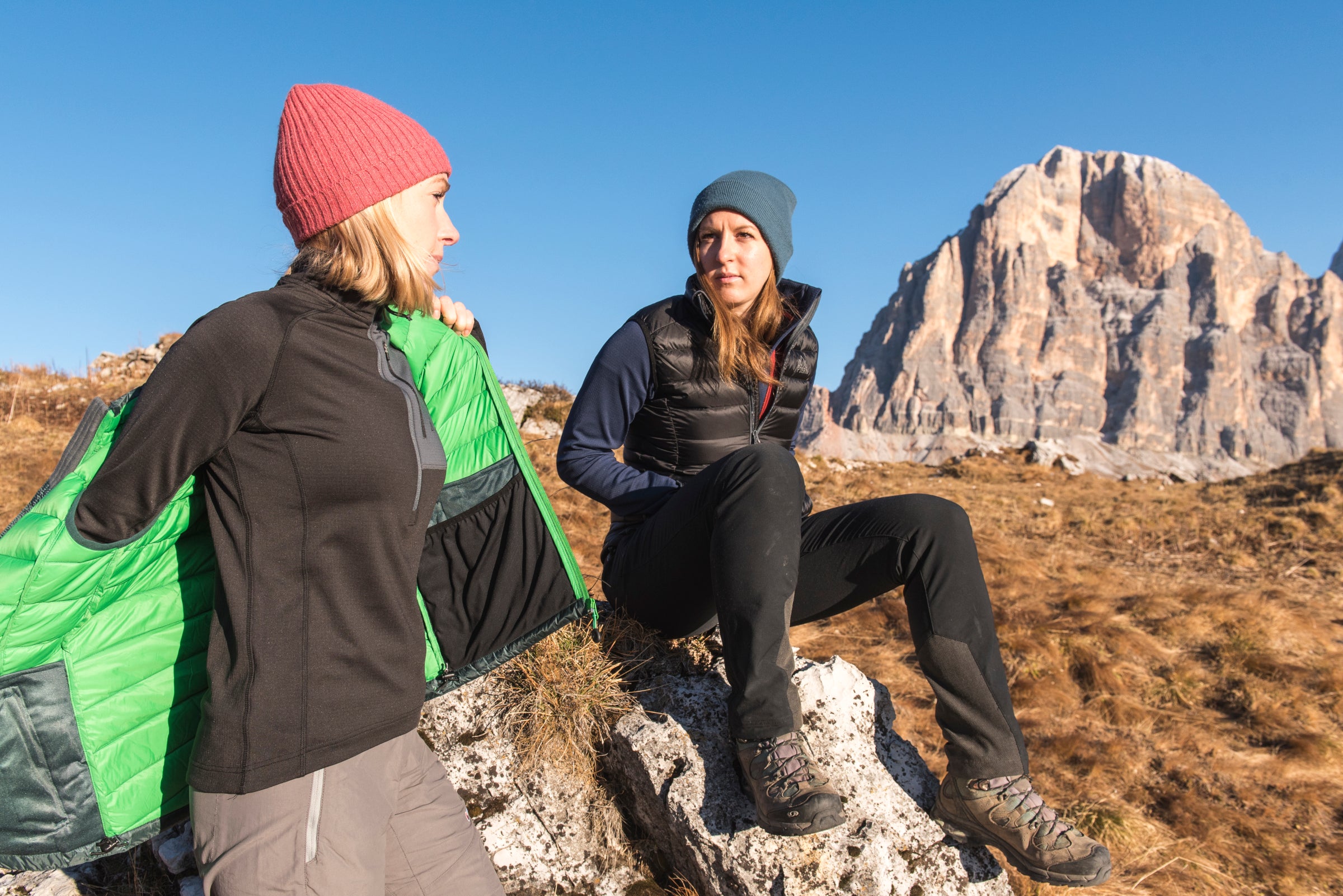 Two women in mid-layers sitting on rocks in a sunny mountain landscape