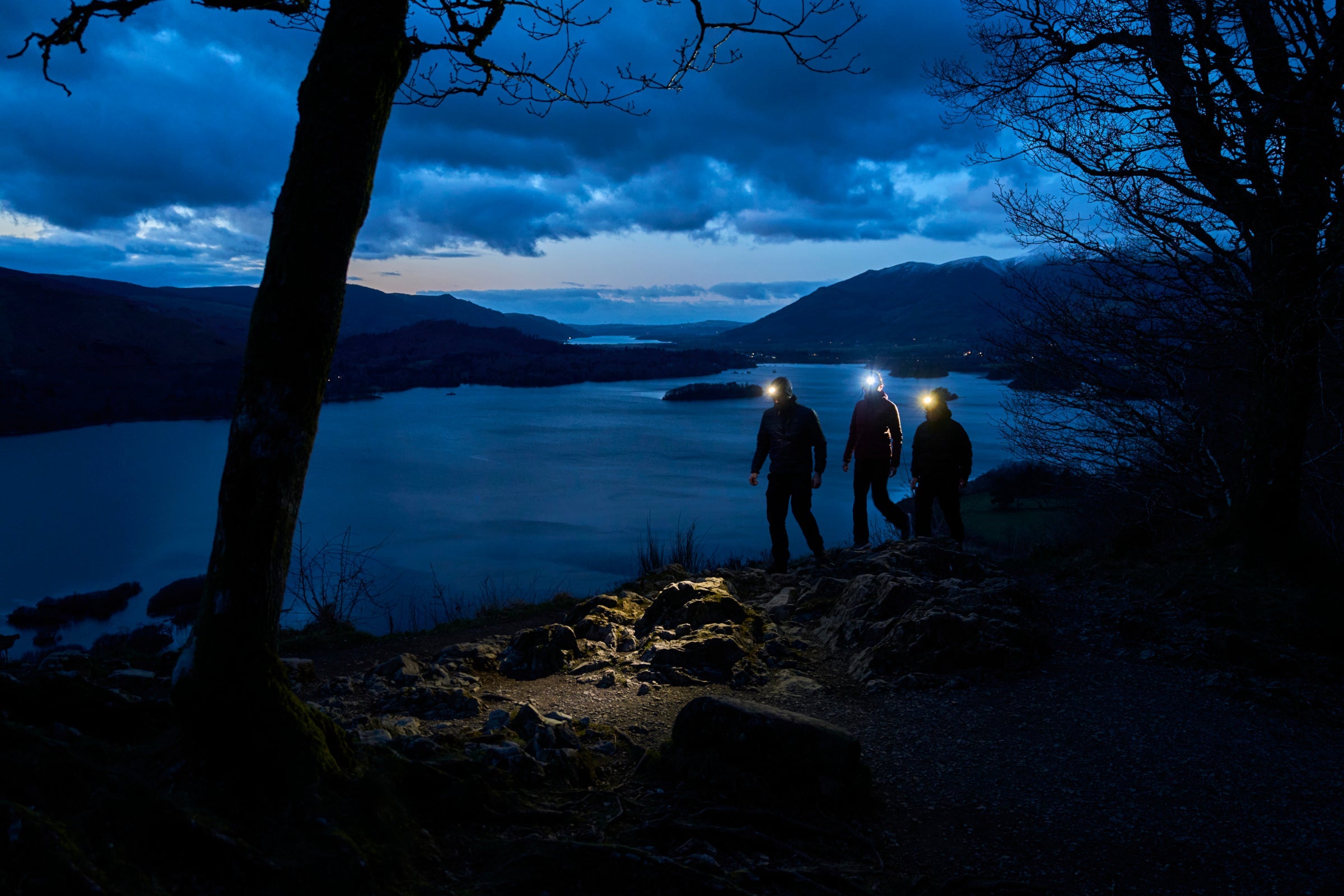 Three hikers wearing headtorches walk along a rocky trail at dusk, overlooking a lake and mountains