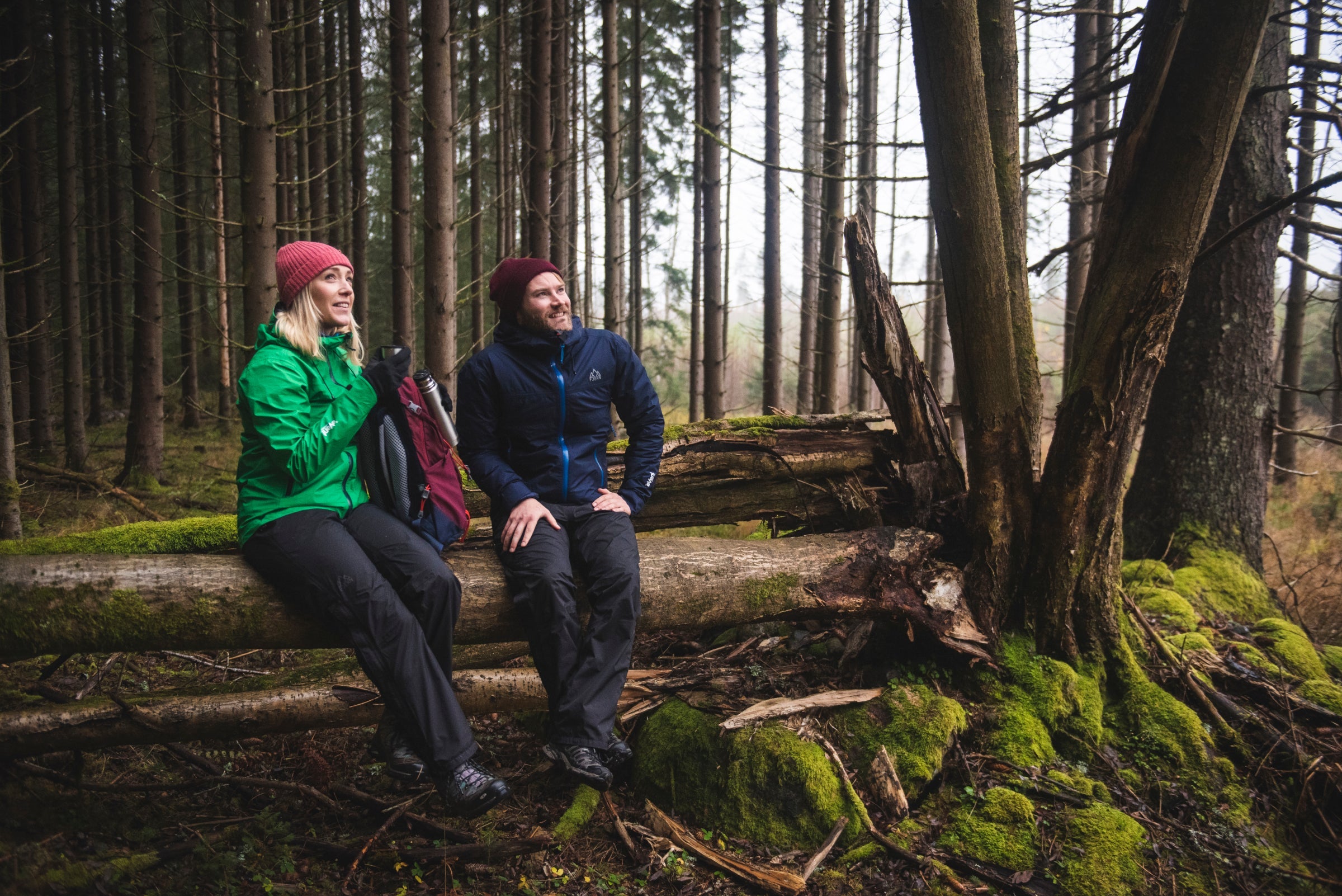 Two people in Fjern clothing sitting on a mossy log in a forest