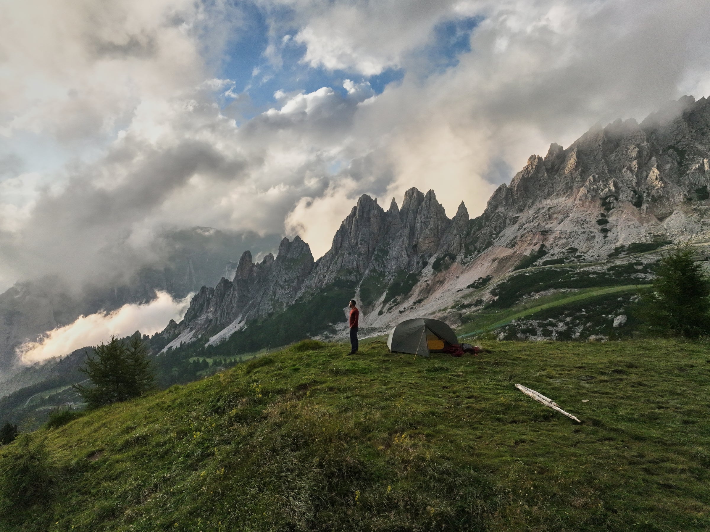 A camper in Fjern gear standing beside a Fjern tent, overlooking dramatic mountain peaks under a cloudy sky