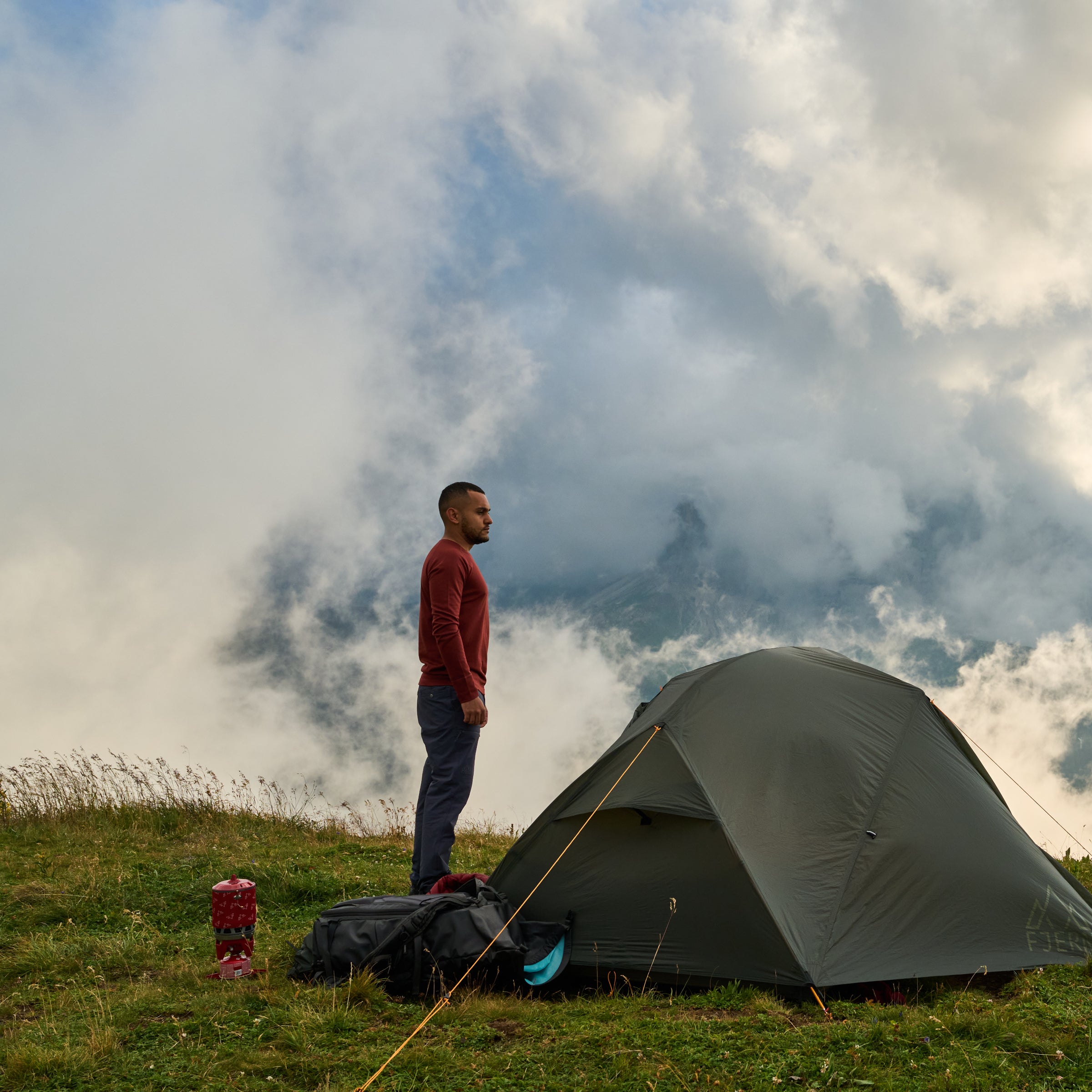 A person, wearing Fjern clothing, standing on a mountain peak, with a Fjern Gokotta Tent pitched overlooking a cloudy valley
