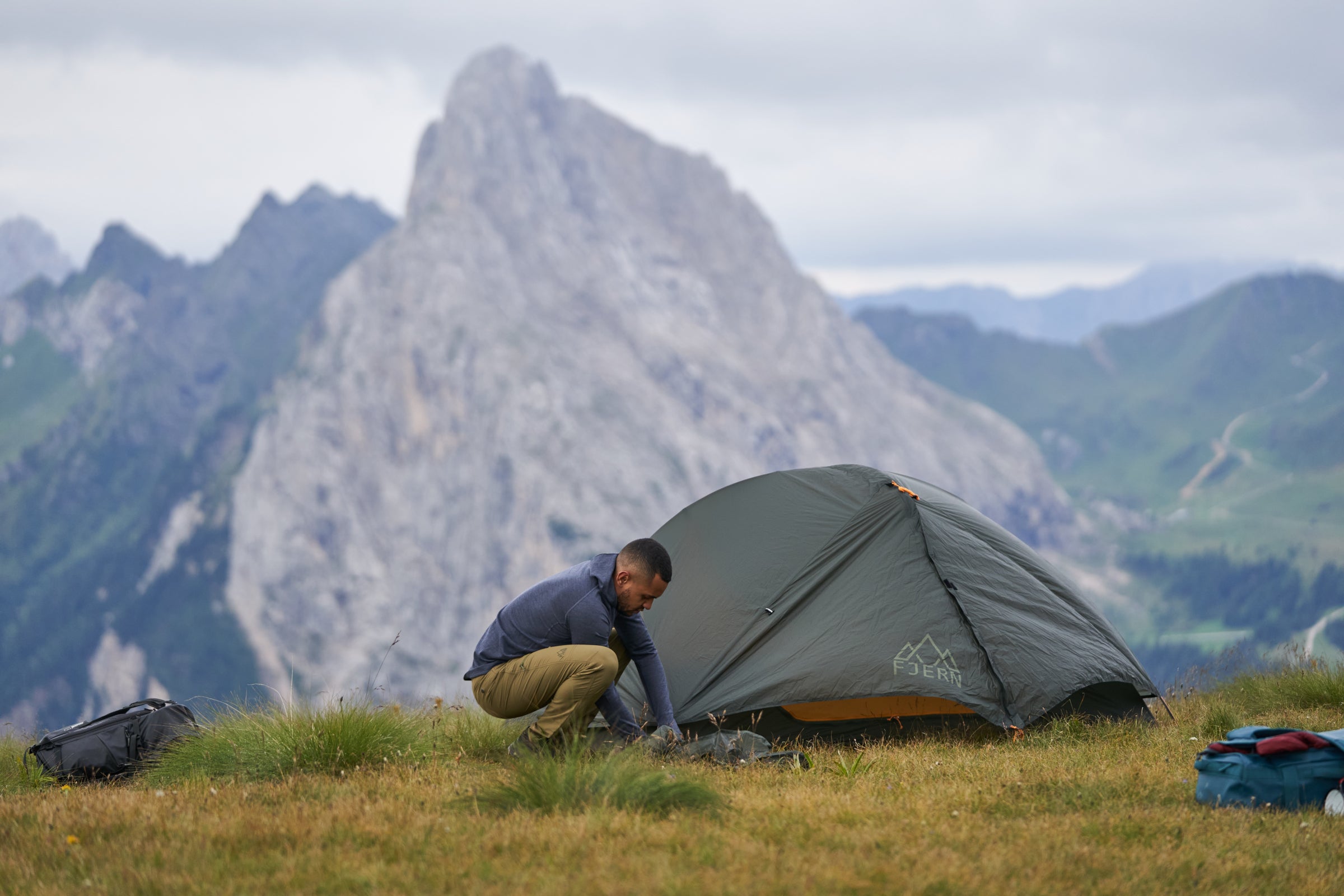 A person setting up a Fjern tent on a grassy mountain ridge with rocky peaks in the background