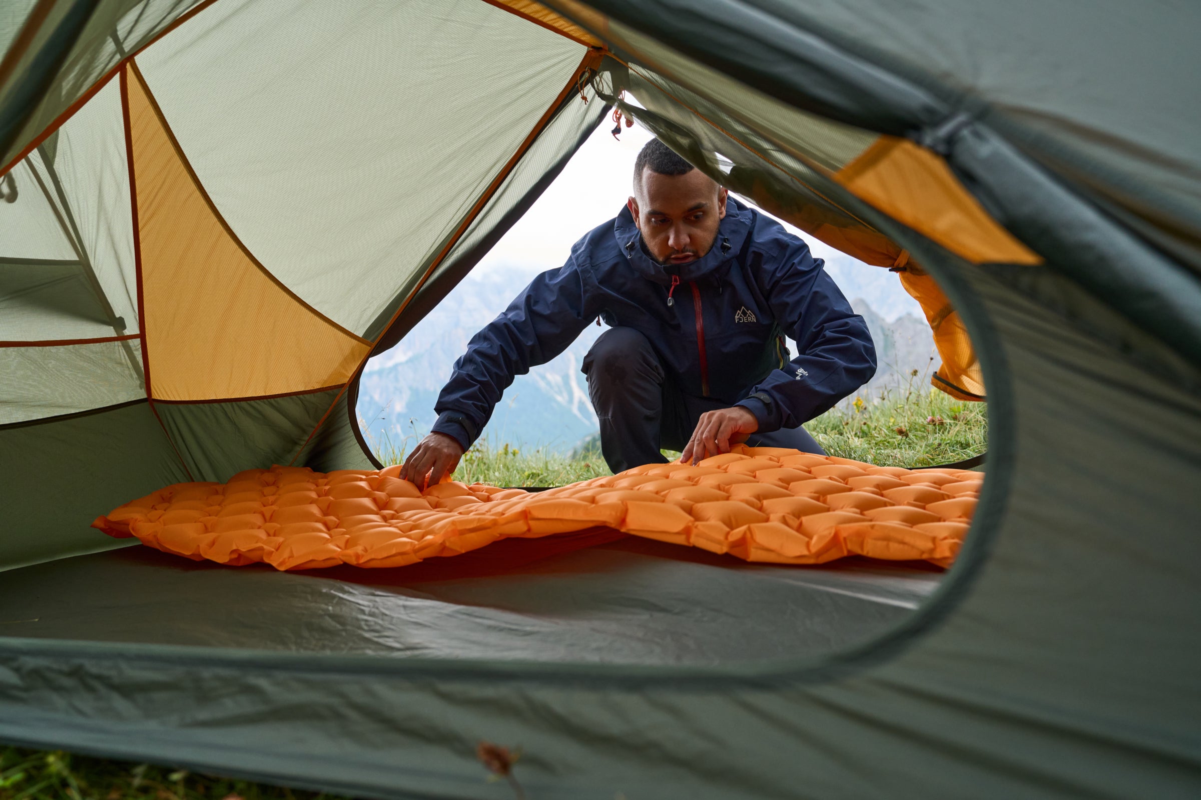 A person arranging an orange sleeping mat inside a Fjern tent with an open door, mountains visible outside