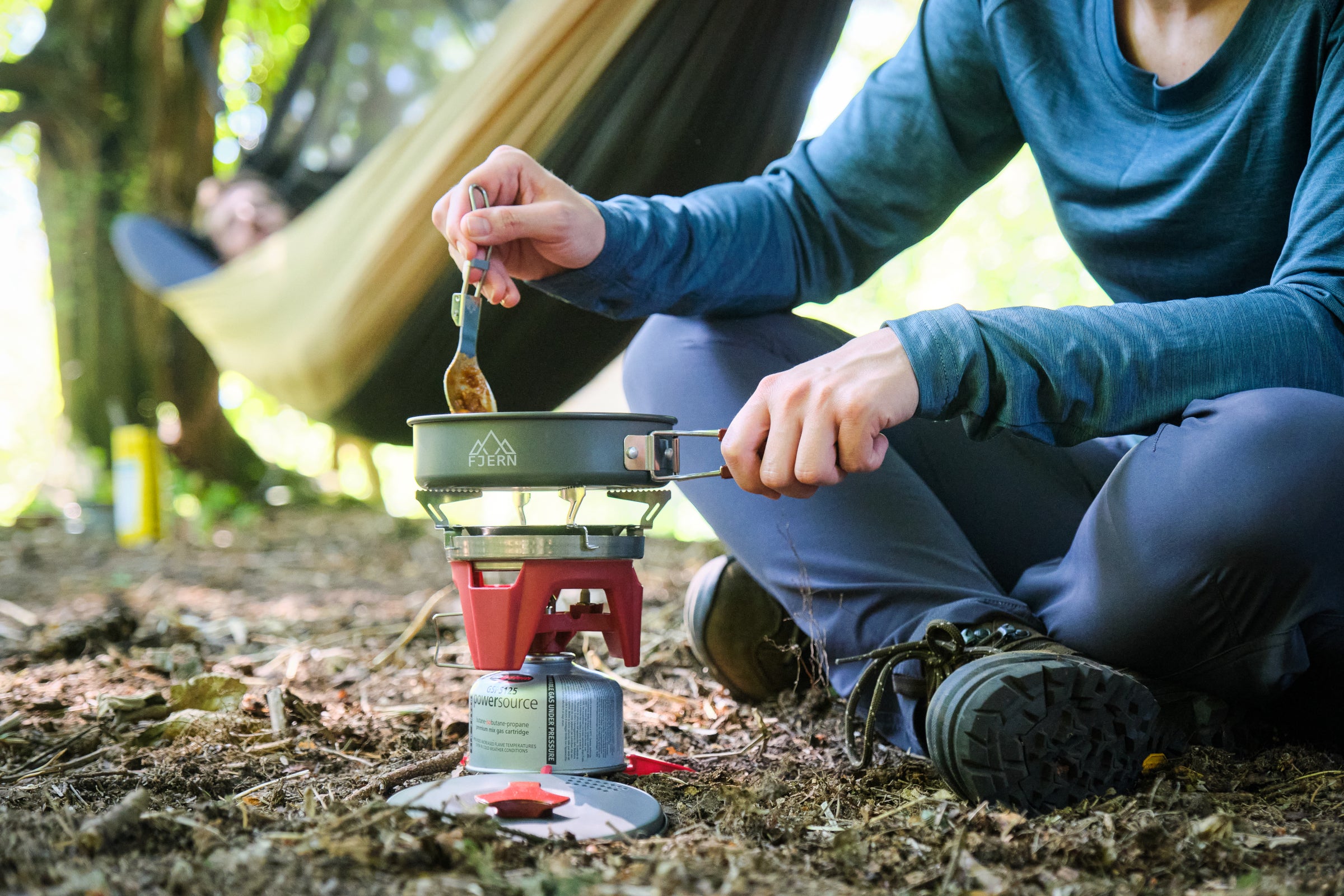 A person cooking on a Fjern portable stove in the forest, with a hammock and trees in the background