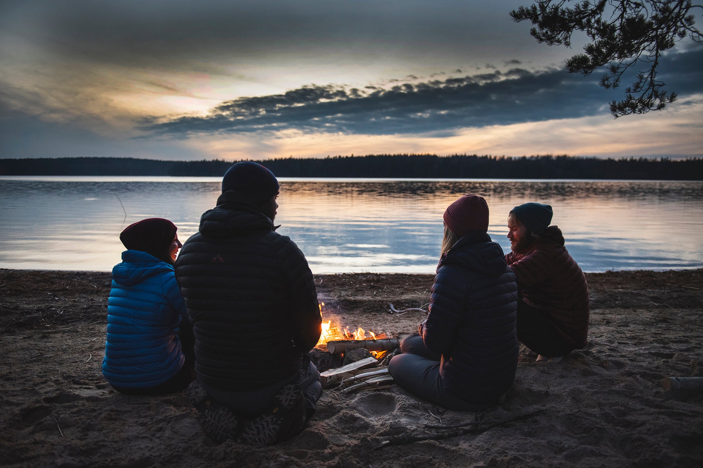 Four people sitting around a campfire on a lakeshore at dusk