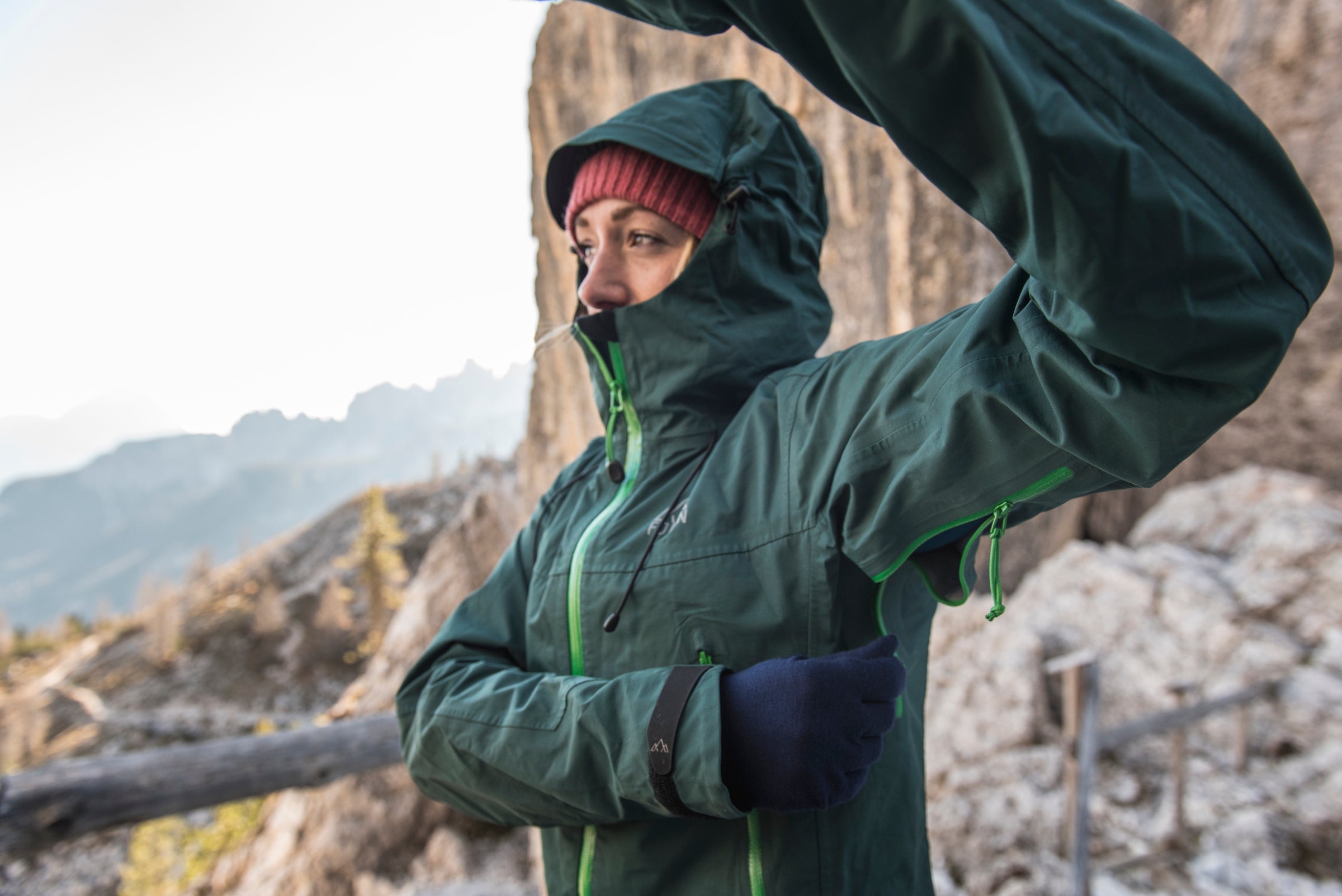 Woman in a green hooded jacket adjusting her armpit zipper in a rocky mountain setting