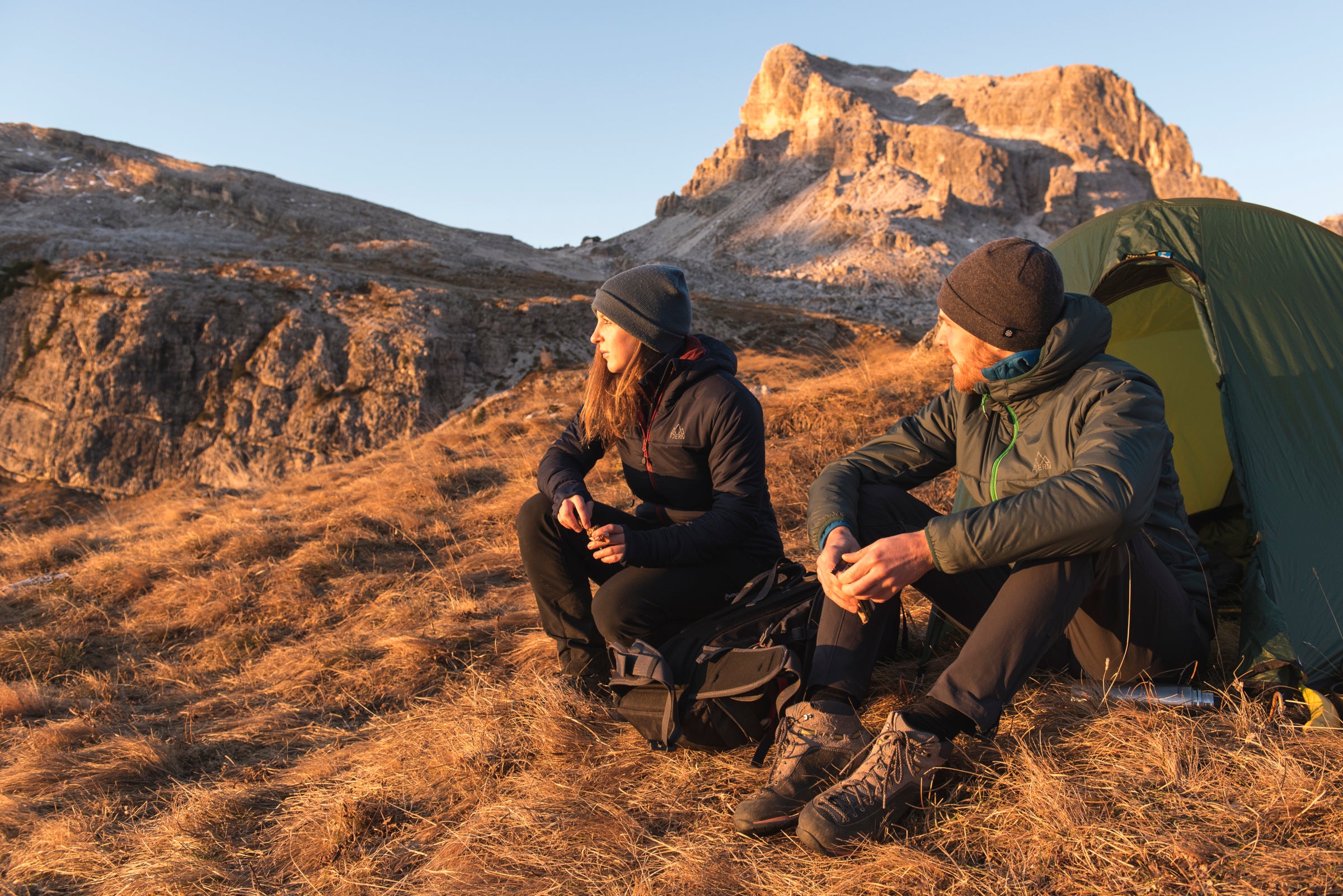 Two hikers sitting near a tent at sunset, with mountains in the background