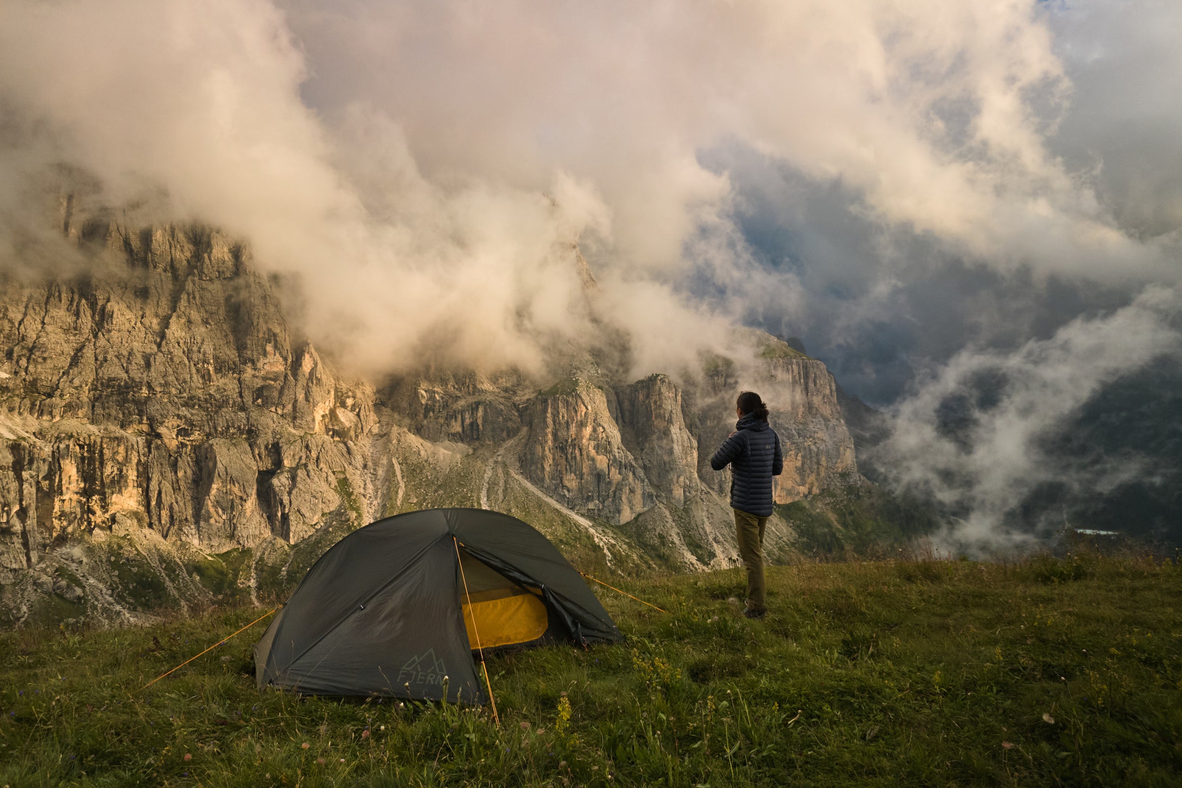 Fjern Gökotta Tent bathed in the soft glow of sunlight, silhouetted against a Dolomite cliff and a sea of churning clouds below