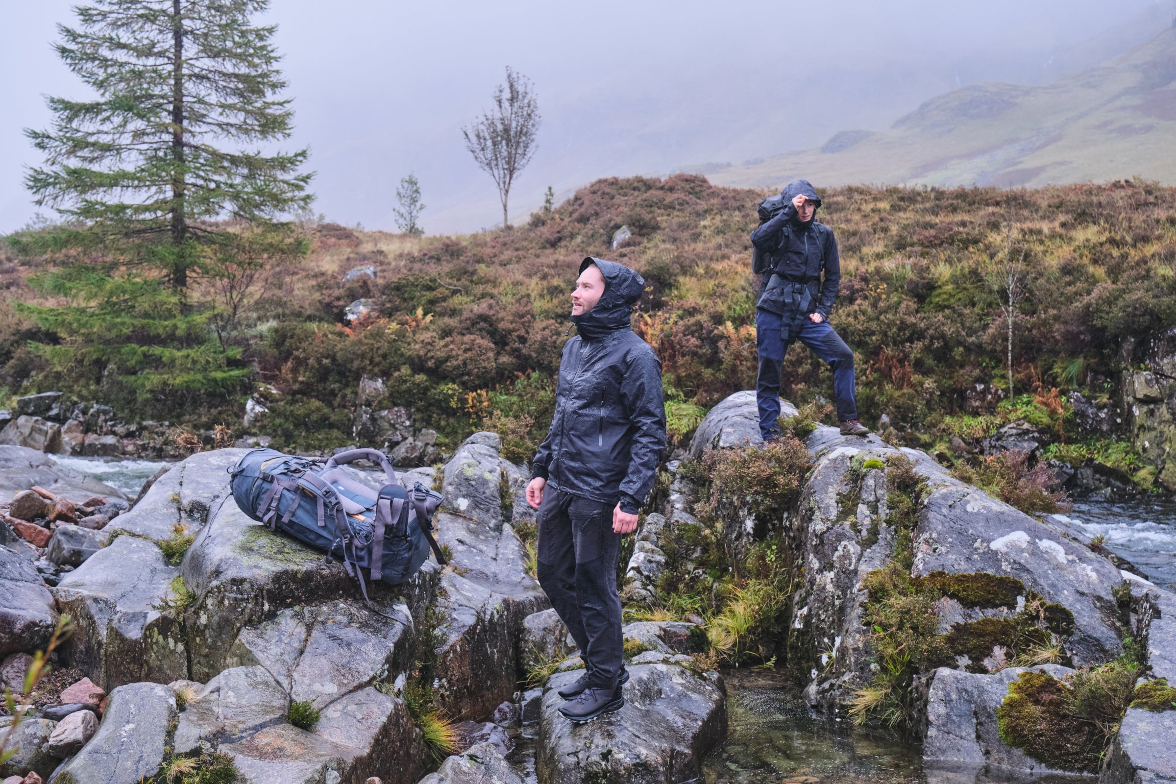 Two hikers in Fjern Orkan waterproof jackets standing on rocks beside a stream in a foggy, mountainous area