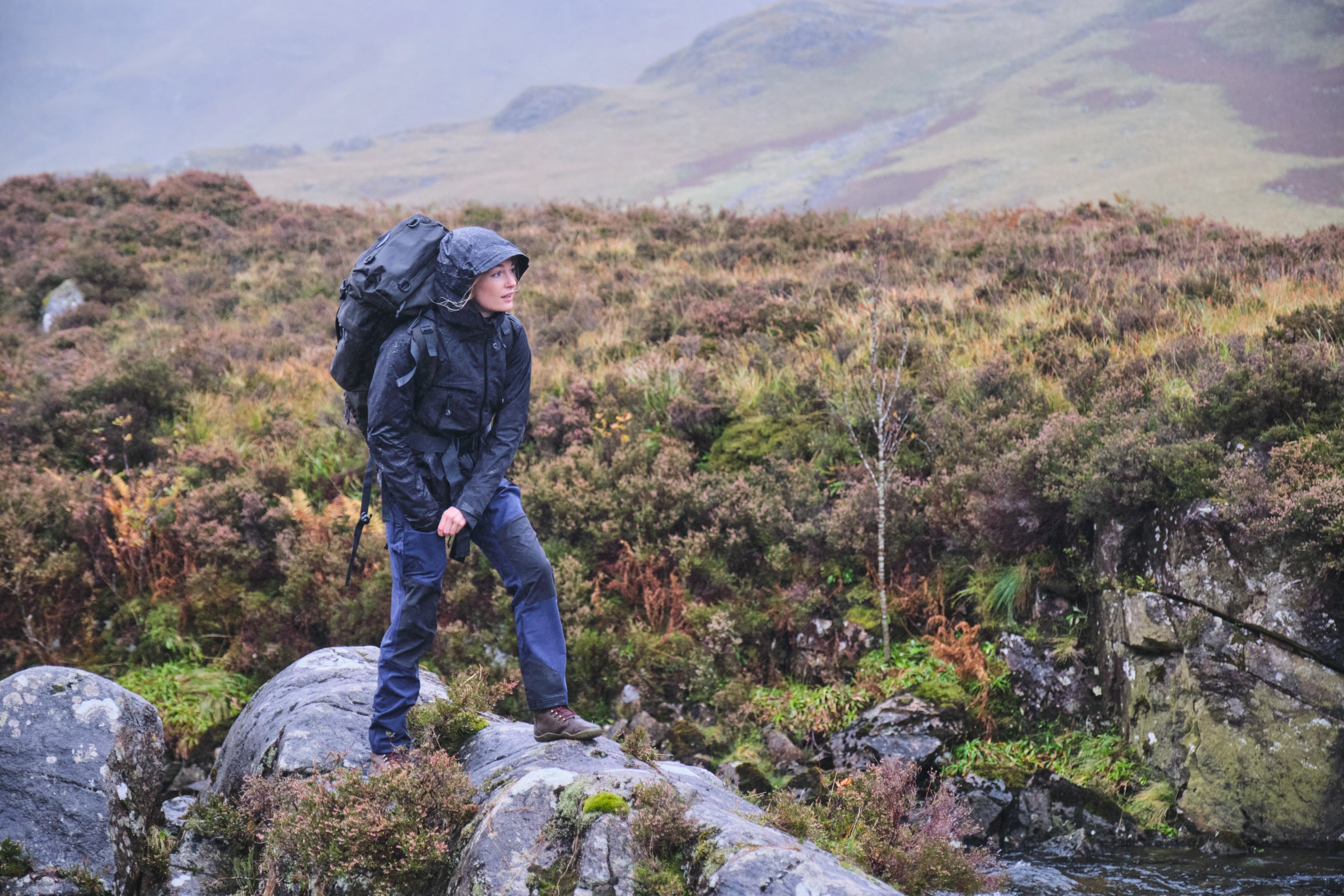 A hiker wearing a Fjern Orkan waterproof jacket standing on a rock in a misty, rugged landscape